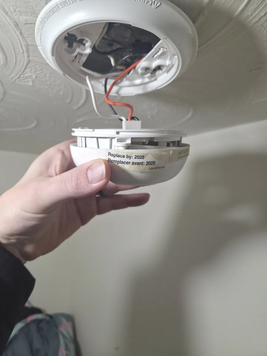 Hand holding a ceiling-mounted smoke alarm beside its replacement-date label.