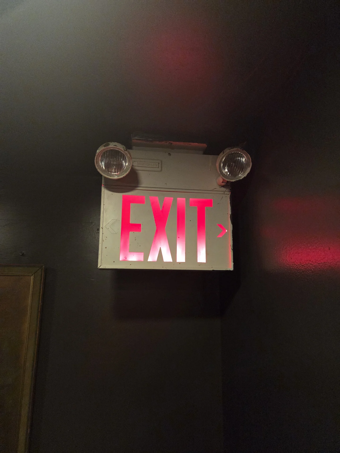 Illuminated EXIT sign with two attached emergency lamp heads on a dark wall.