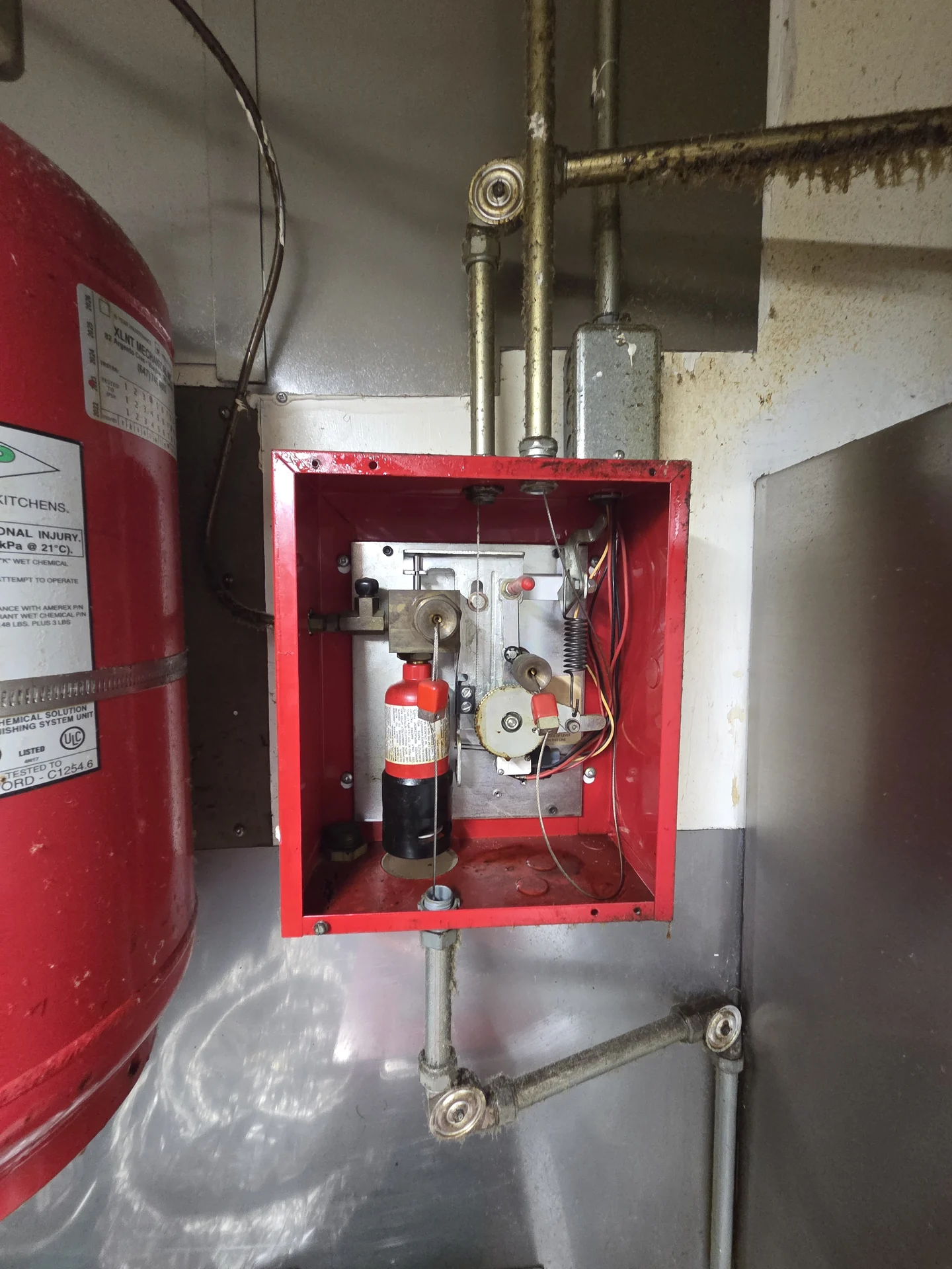 Open red kitchen suppression release enclosure beside a suppression cylinder and metal conduit.